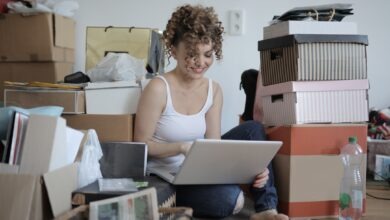 Smiling female customer in casual wear sitting on floor in untidy room with stacks of cardboard boxes and making orders in online store while using laptop