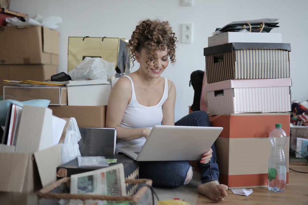 Smiling female customer in casual wear sitting on floor in untidy room with stacks of cardboard boxes and making orders in online store while using laptop